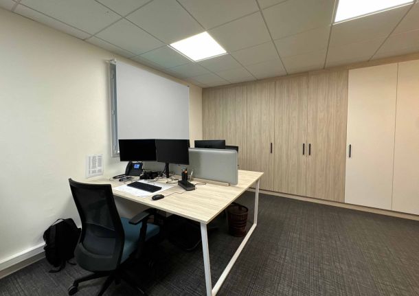 Bureau professionnel aménagé sur mesure avec une grande armoire en bois clair et blanc, un bureau en bois clair équipé d'écrans doubles, et un store bleu apportant une touche de couleur à un espace de travail moderne.