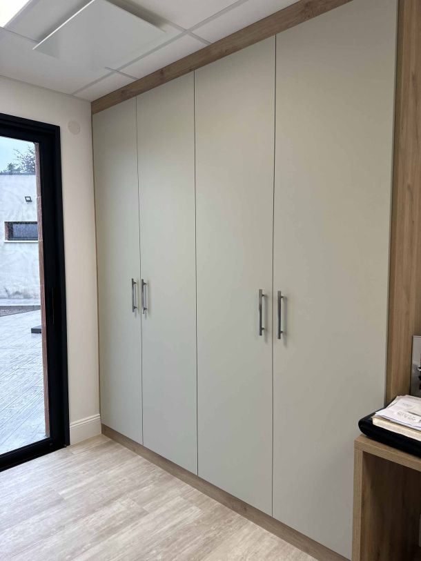 Placard sur mesure avec portes battantes en finition claire et encadrement en bois, installé dans une pièce lumineuse avec un sol en parquet et une porte-fenêtre donnant sur l'extérieur.