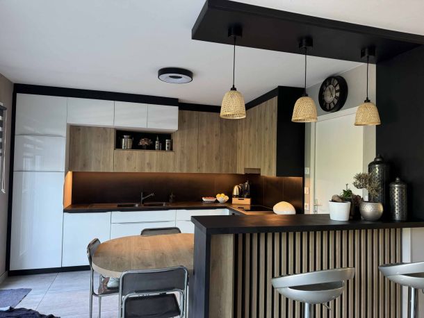 Cuisine moderne avec façade blanche laquée, éléments en bois clair, plan de travail sombre, éclairage LED, bar à lattes en bois, suspensions en osier et table à manger ronde avec chaises métalliques.