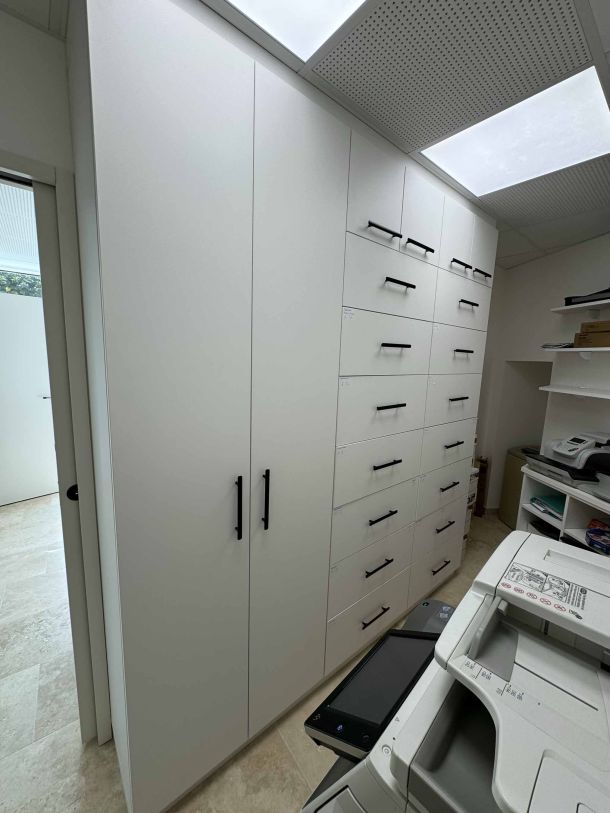 Meuble d'archives sur mesure avec armoires blanches et tiroirs équipés de poignées noires, intégré dans un espace professionnel avec plafond lumineux et imprimante multifonction.